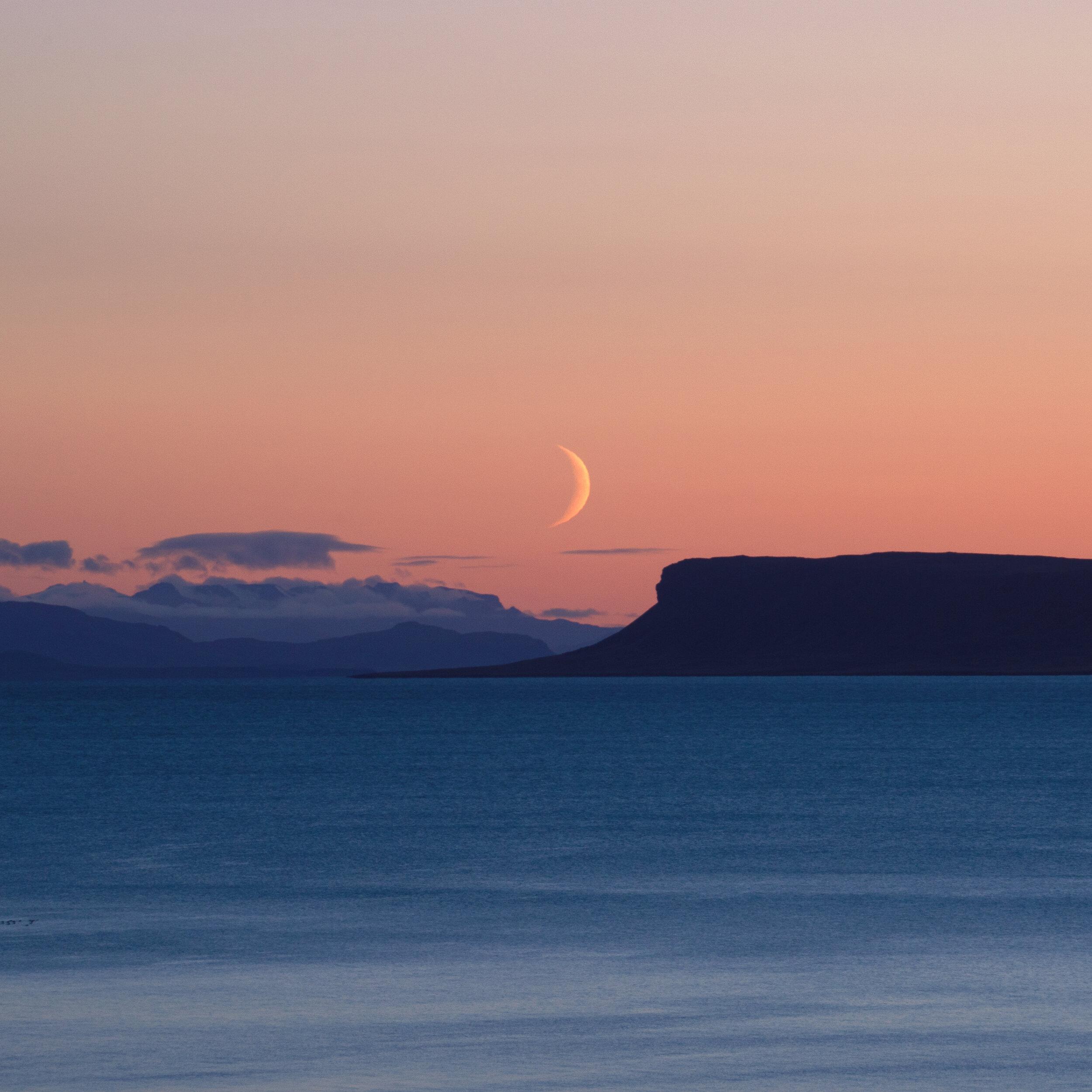 A crescent moon setting over dark cliffs and the sea at dusk
